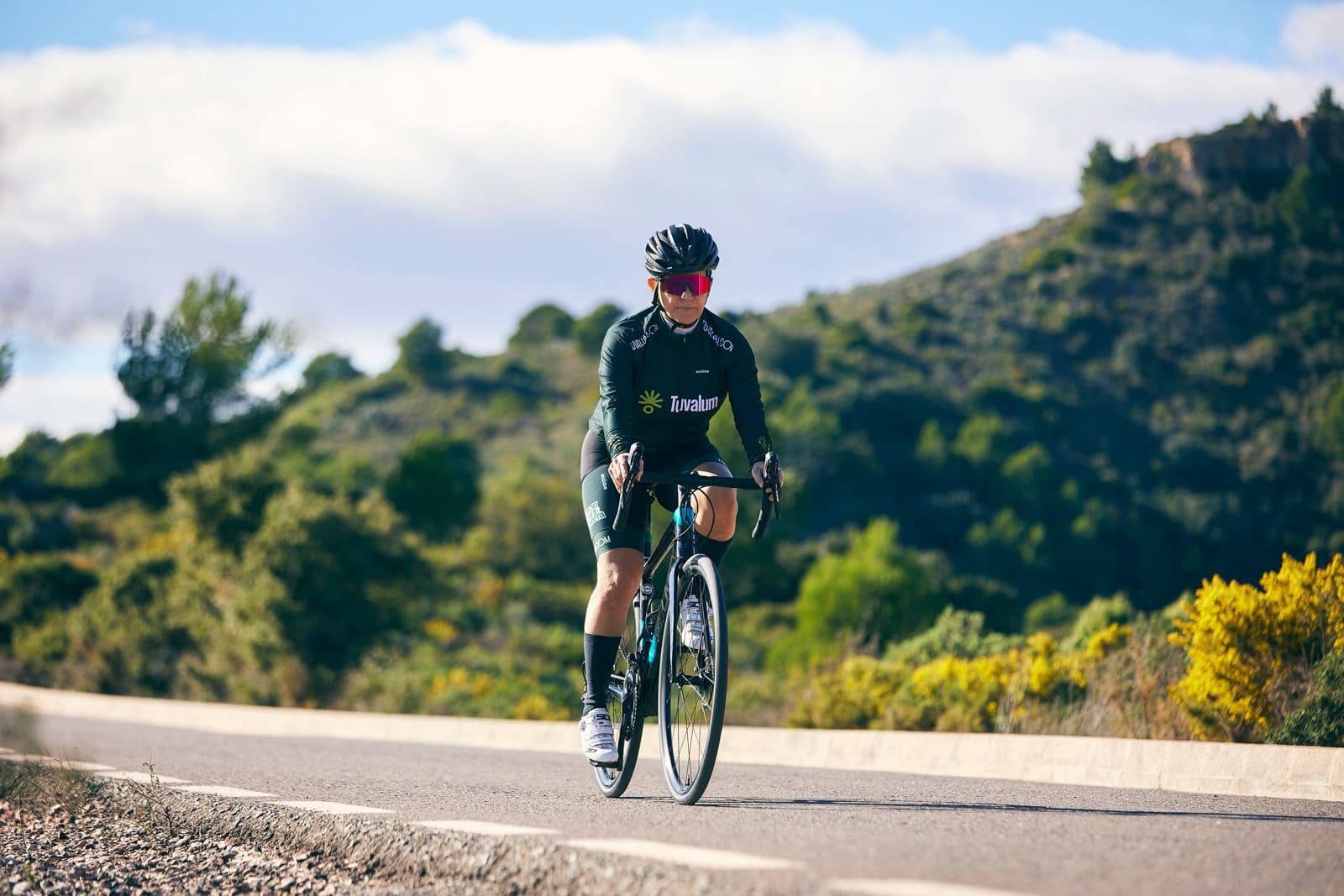 Cyclist riding a road bike on a scenic paved road surrounded by green hills and clear blue sky.