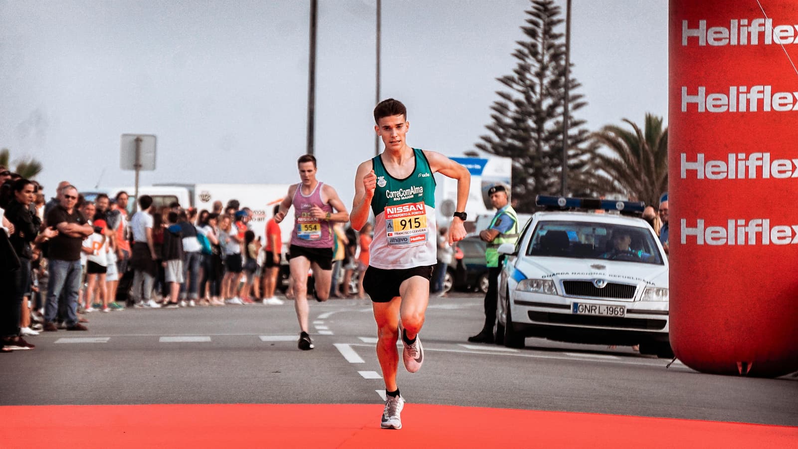 A focused triathlete runs along the race course during a triathlon, wearing a race bib and athletic gear, with other competitors and spectators visible in the background.