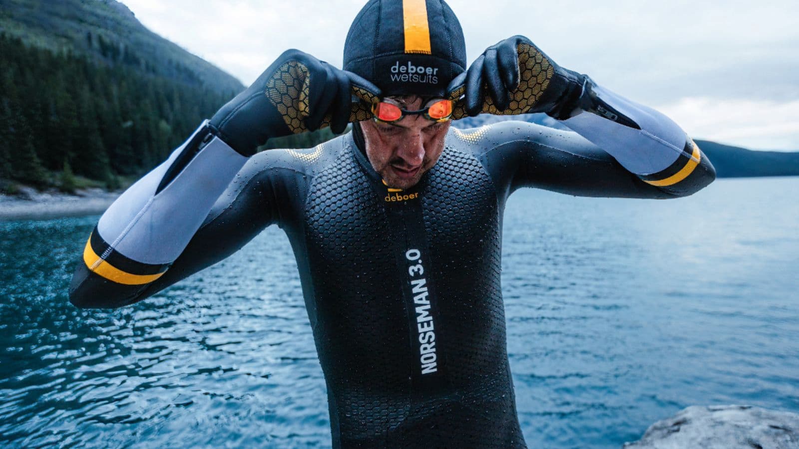 Triathletes wearing wetsuits and goggles preparing at the water's edge before an open water swim start.
